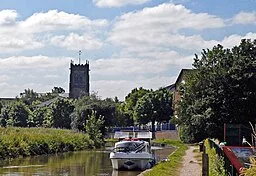The parish church of St. Michael and All Angels, in Middlewich is shown in the background seen over the Trent and Mersey Canal. A boat is moored on the canal