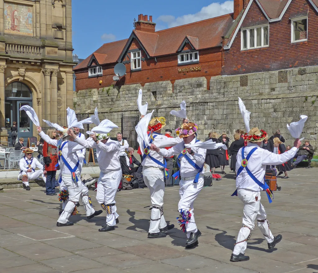 Morris dancers dancing. They are dressed in white wearing blue sashes across their torso. They are holding handkerchiefs
